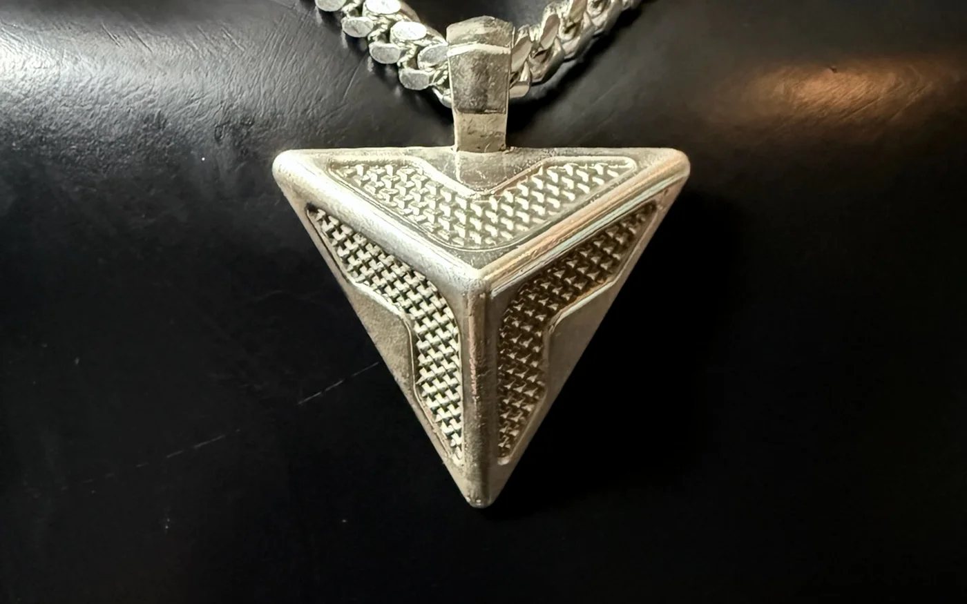 A handmade silver triangular pendant on a heavy chain, lit against black leather